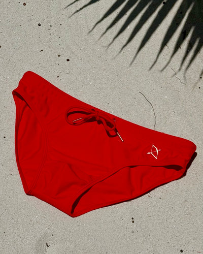 Apollo Swim Brief in Chilli Red flat lay on sand, showing gold metal Archer hardware and drawcord